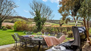 An outdoor patio with a table set for dining, surrounded by chairs, a grill, and a view of rolling hills. - 8 Bed in Dorchester (oc-93723) (Minterne Magna)