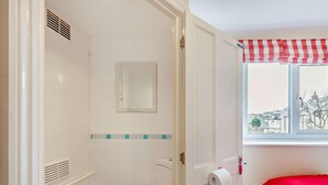 Een badkamer met toilet, spiegel, raam met rood-wit geruit gordijn en uitzicht op huizen.