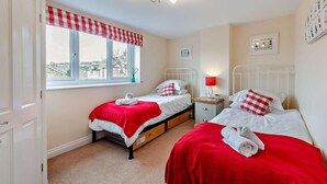 Een slaapkamer met twee bedden, een raam met een rood-wit geruit gordijn, een nachtkastje met een lamp en een wandklok.
