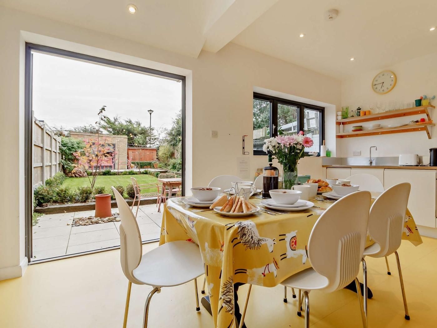 A dining area with a table set for four, chairs, a window with a view of the garden, and a kitchen area with shelves.