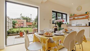 A dining area with a table set for four, chairs, a window with a view of the garden, and a kitchen area with shelves.