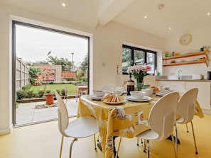 Een eetruimte met een tafel voor vier personen, stoelen, een raam met uitzicht op de tuin en een keuken met schappen.