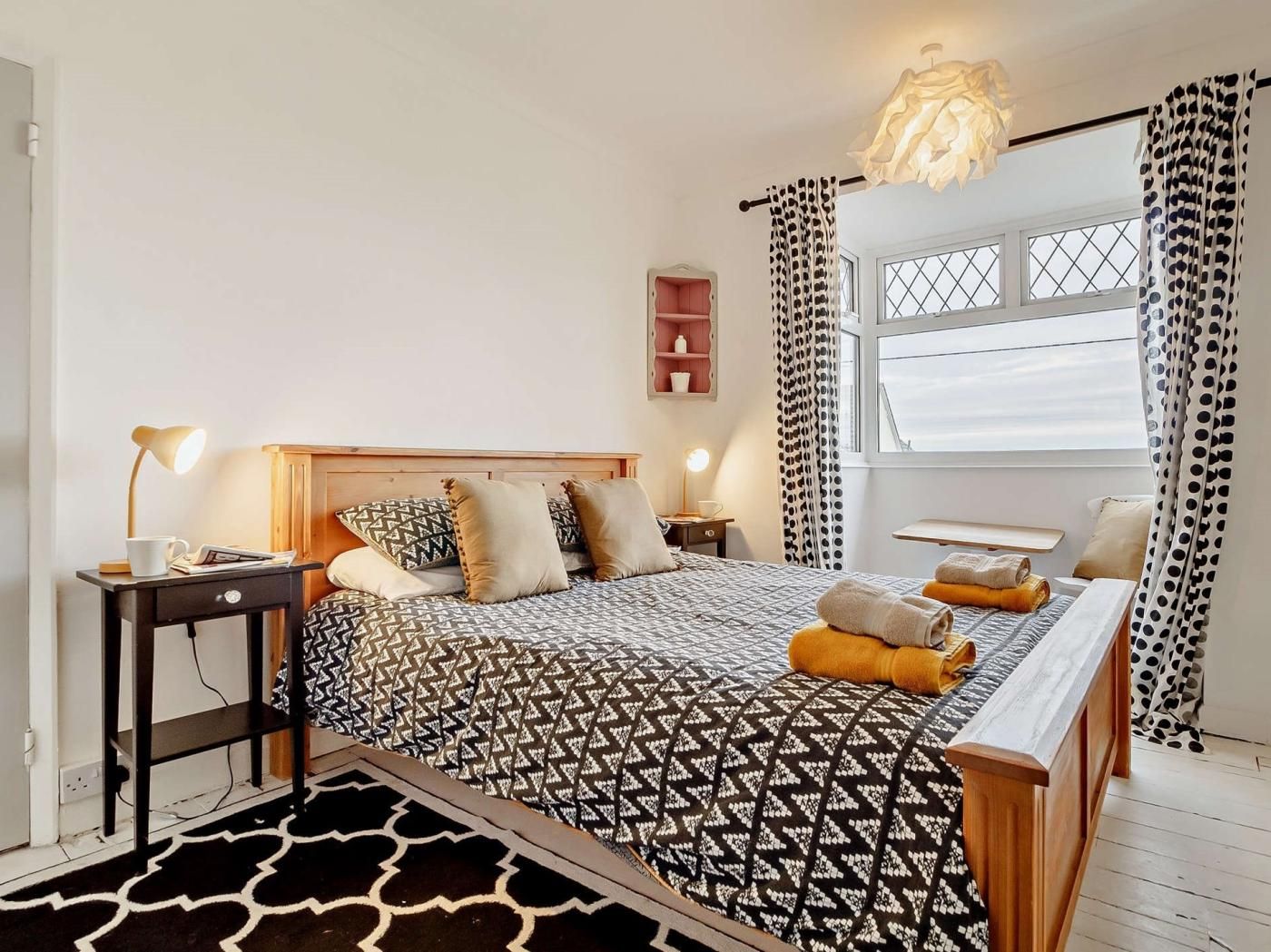 A bedroom with a bed, bedside tables, a desk, a chair, a window, and a hanging light fixture.
