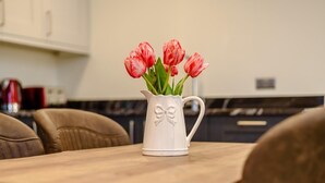 Un pichet blanc orné d’un motif en forme de nœud, contenant des tulipes roses, est posé sur une table en bois dans une cuisine.