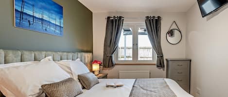 Habitación de hotel con cama, un cuadro, ventana con cortinas, mesita de noche con lámpara y televisor montado en la pared.