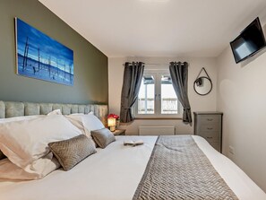 Habitación de hotel con cama, un cuadro, ventana con cortinas, mesita de noche con lámpara y televisor montado en la pared.