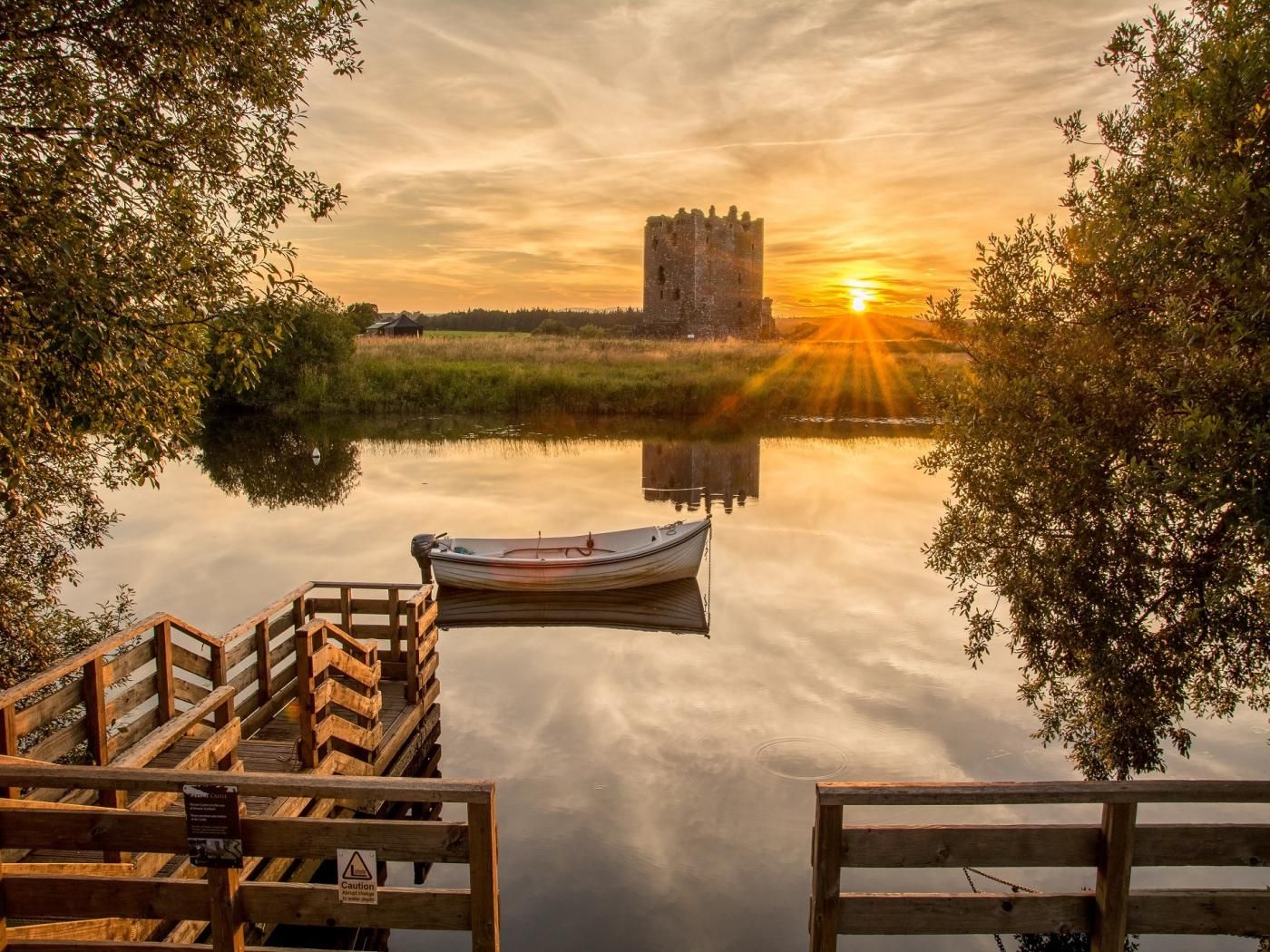 A calm body of water with a wooden dock, a boat, and a castle in the distance during sunset.