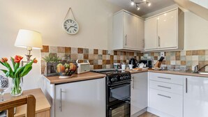 Een kleine keuken met witte kastjes, een zwarte oven, een magnetron en een kookplaat. Er hangt een klok aan de muur, op het aanrecht staat een vaas met bloemen en een lamp.