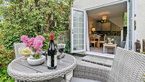 Een terras met rieten meubilair, een tafel met wijn en bloemen, en uitzicht op een keuken door een geopende deur.