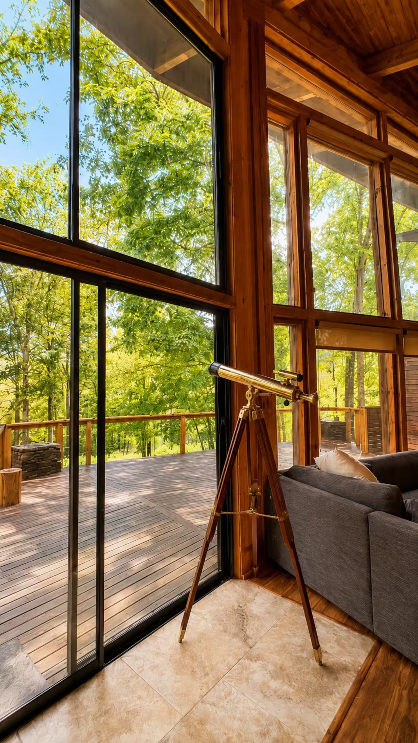 Una habitación con ventanas amplias, un telescopio, una terraza de madera y un sofá gris.