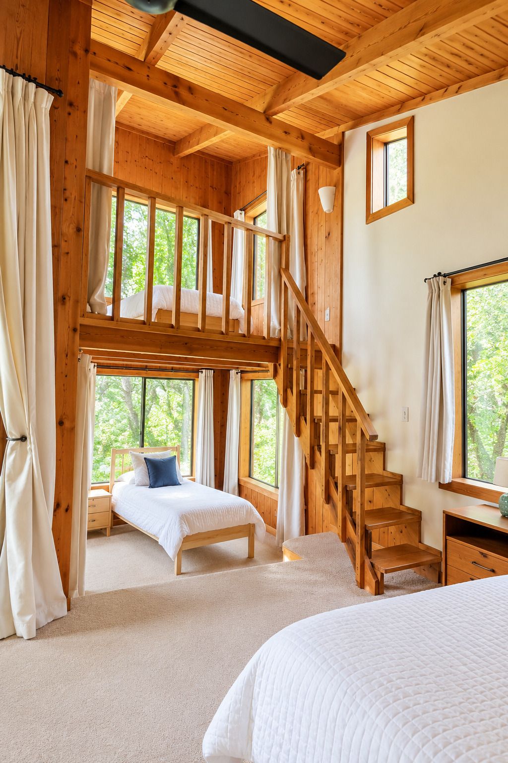 Dormitorio de cabaña de madera con cama de altillo, escalera y ventanas amplias.