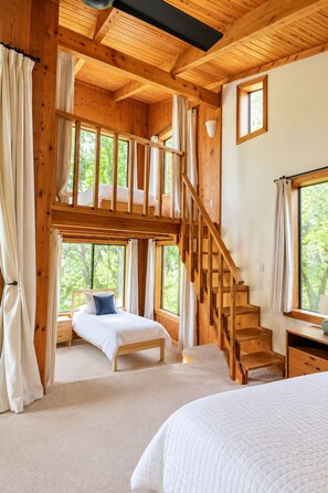 A wooden cabin bedroom with a loft bed, a staircase, and large windows.