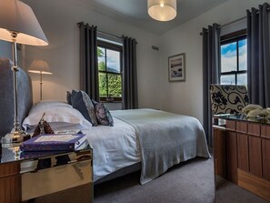 Een slaapkamer met een bed, nachtlampen, een nachtkastje met lamp, een dressoir en een raam met gordijnen.