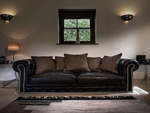 Ein schwarzes Ledersofa mit mehreren Kissen, ein Fenster mit Blick auf Grünflächen und zwei Wandleuchten.