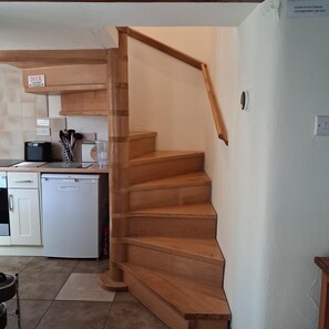 A wooden staircase leading to a landing with a kitchen area in the background. - 2 Bed in Ashford In The Water (oc-85823) (Ashford in the Water)