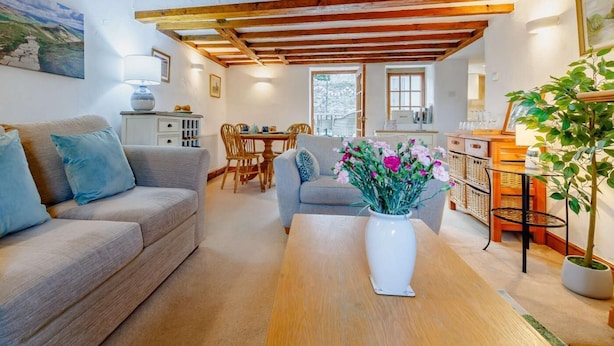 A cozy living room with a sofa, a coffee table, and a vase of flowers. There is a dining area with a table and chairs, and a kitchen area with a counter and a plant. - 2 Bed in Ashford In The Water (oc-85823) (Ashford in the Water)