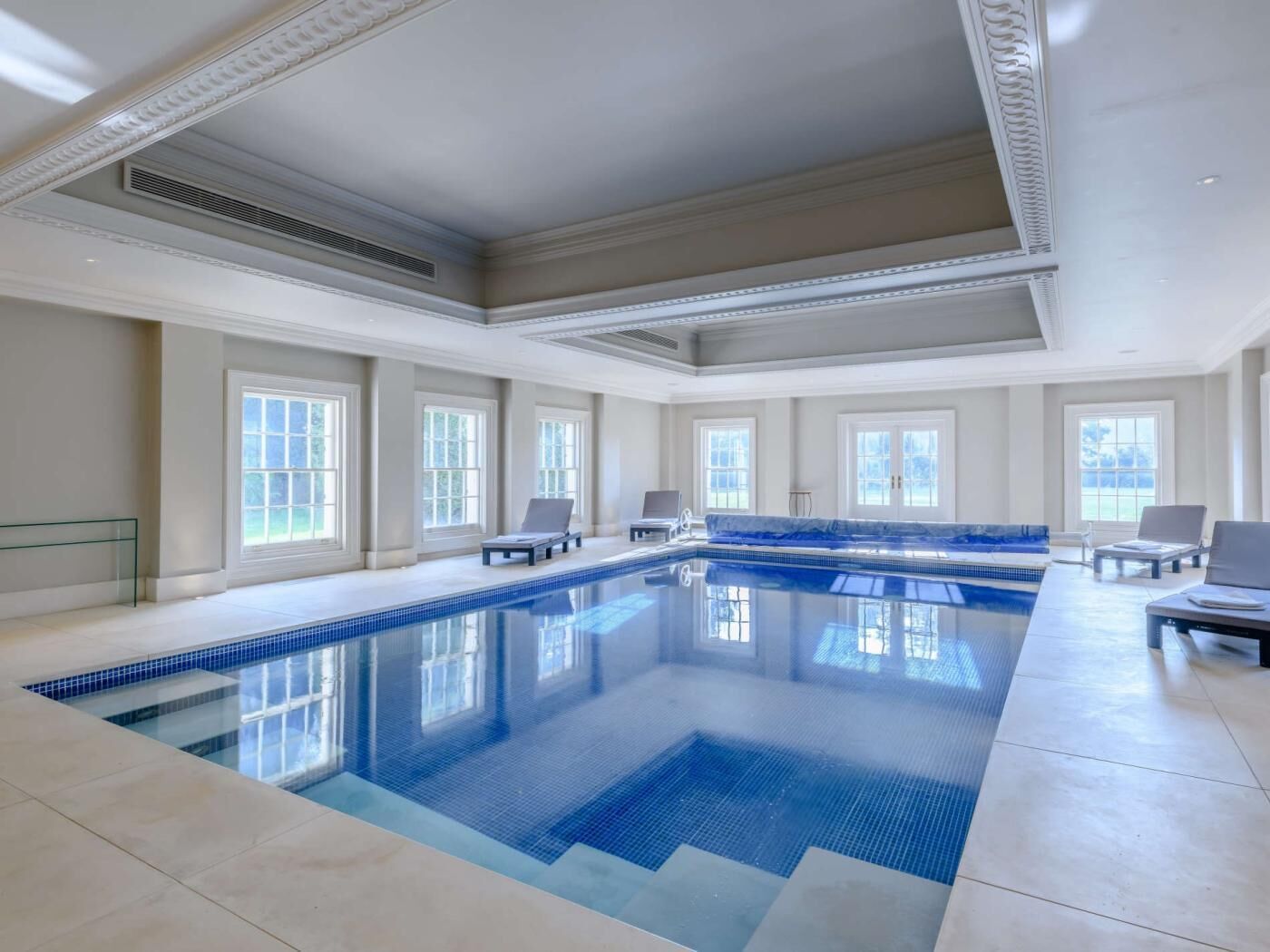 A large indoor swimming pool with clear blue water, surrounded by lounge chairs and large windows.