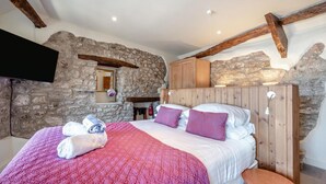 A bedroom with a stone wall, wooden beams, a bed with pink bedding, a wooden wardrobe, and a flat-screen TV mounted on the wall. - 3 Bed in Tenby (oc-fb225) (Tenby)