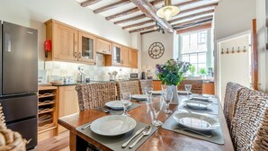 A kitchen with wooden cabinets, a stainless steel refrigerator, and a dining area with wicker chairs and a table set for four. - 3 Bed in Tenby (oc-fb225) (Tenby)