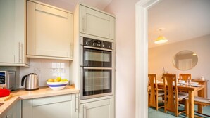 A compact kitchen with a kettle, a bowl of fruit, and a double oven. - 3 Bed in Keswick (oc-sz316) (Keswick)