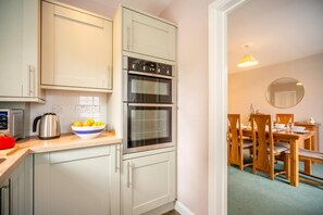 A compact kitchen with a kettle, a bowl of fruit, and a double oven. - 3 Bed in Keswick (oc-sz316) (Keswick)