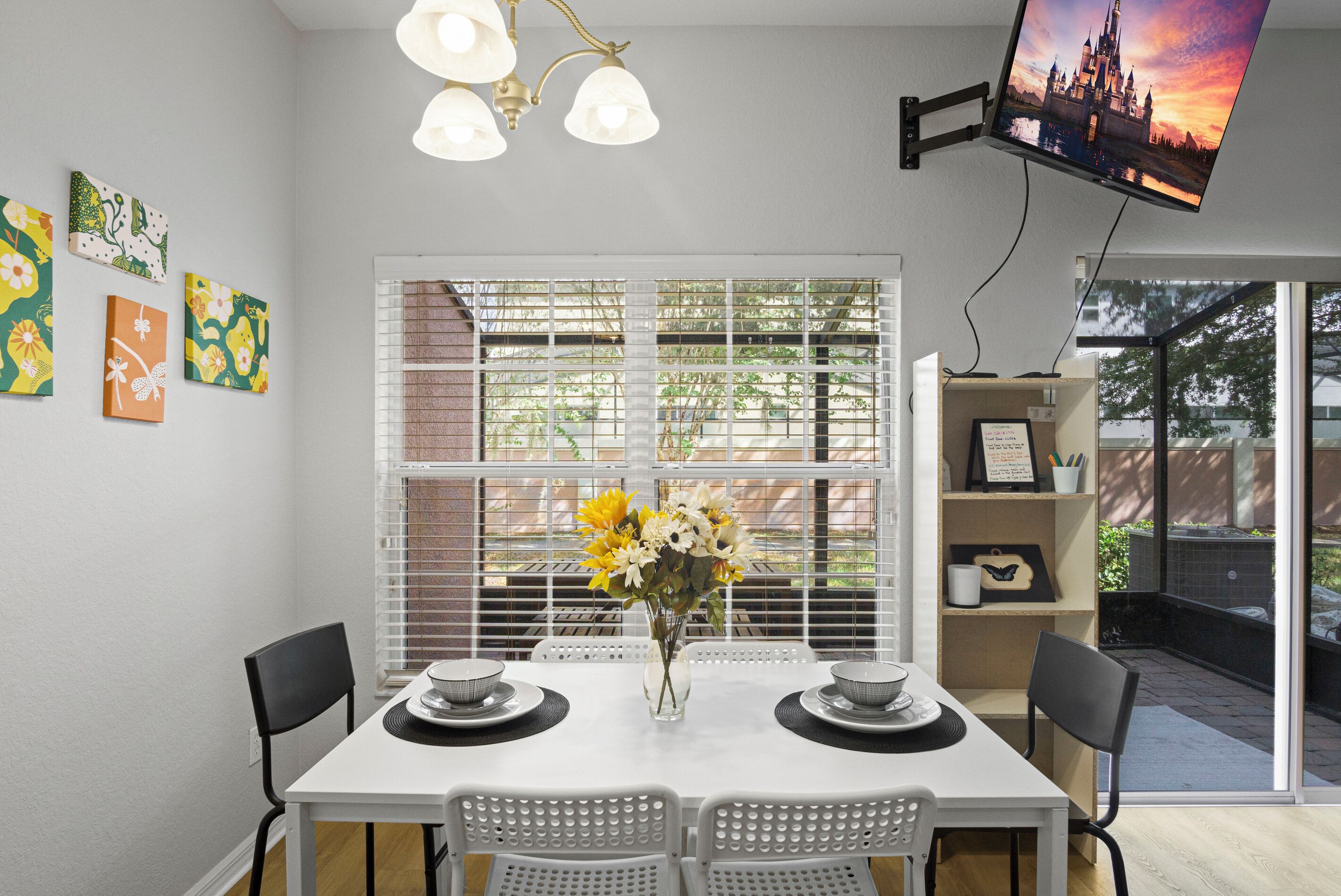 A dining area with a white table, chairs, a vase with flowers, and a TV mounted on the wall.