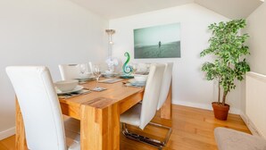 A dining area with a wooden table set for six, white chairs, a potted plant, and a framed picture on the wall.