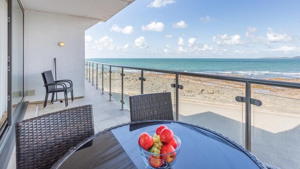 A balcony with a glass railing overlooking a beach, a table with a fruit bowl, and two wicker chairs.