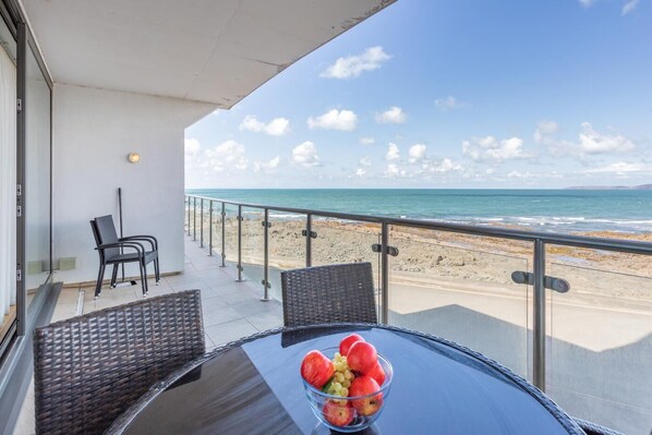 A balcony with a glass railing overlooking a beach, a table with a fruit bowl, and two wicker chairs.