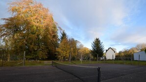 Ein Tennisplatz im Freien mit Netz, umgeben von Bäumen und einem Gebäude im Hintergrund.