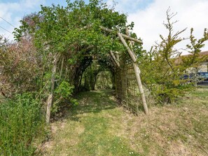 Caminho serpenteando por um jardim com um arco de madeira, cercado por vegetação e um edifício de pedra ao fundo.