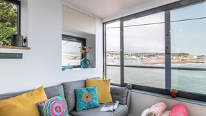 Ein modernes Wohnzimmer mit einem grauen Sofa, bunten Kissen, einem kleinen Tisch mit einer Zeitschrift und einem großen Fenster mit Blick auf die Küste.