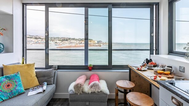 Ein modernes Zimmer mit großem Fenster zum Strand, einer Couch, einem kleinen Tisch und einer Küchenzeile.
