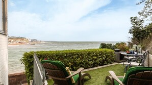 Ein Balkon mit zwei Holzstühlen, einem kleinen Tisch, einer Flasche und Blick auf das Meer und einen Steg.