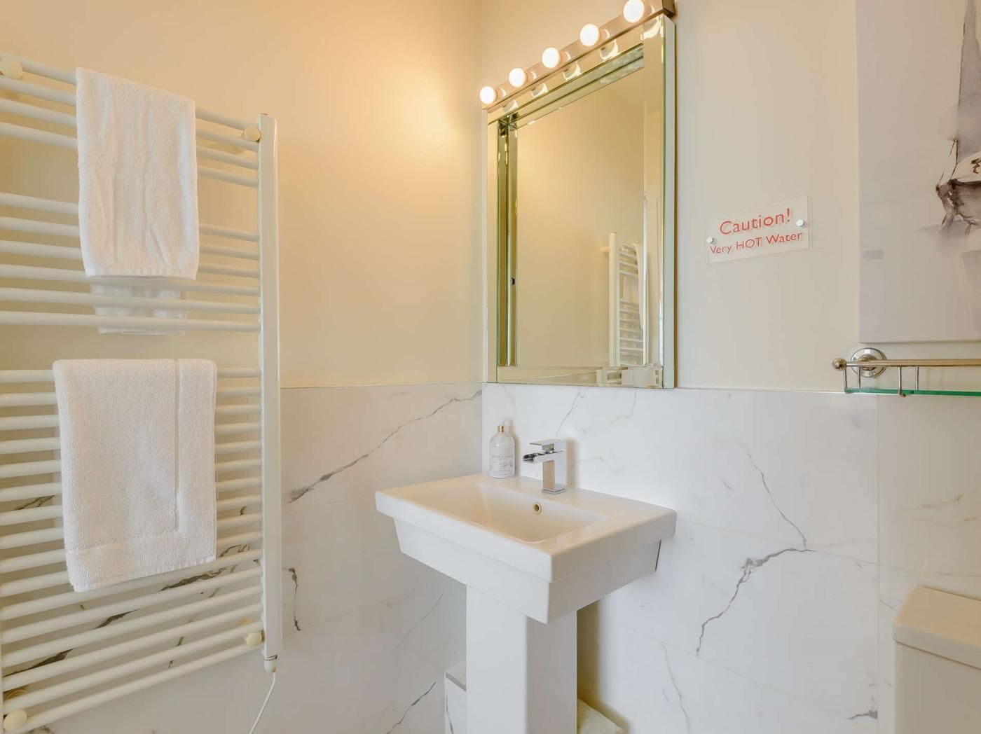 A bathroom with a white sink, a mirror with lights, a towel rack, and a caution sign about hot water.