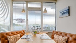A dining area with a wooden table, two brown cushioned benches, and a view of the sea through large windows with blinds. - 1 Bed in Hythe (oc-89969) (Hythe)