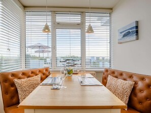 A dining area with a wooden table, two brown cushioned benches, and a view of the sea through large windows with blinds. - 1 Bed in Hythe (oc-89969) (Hythe)