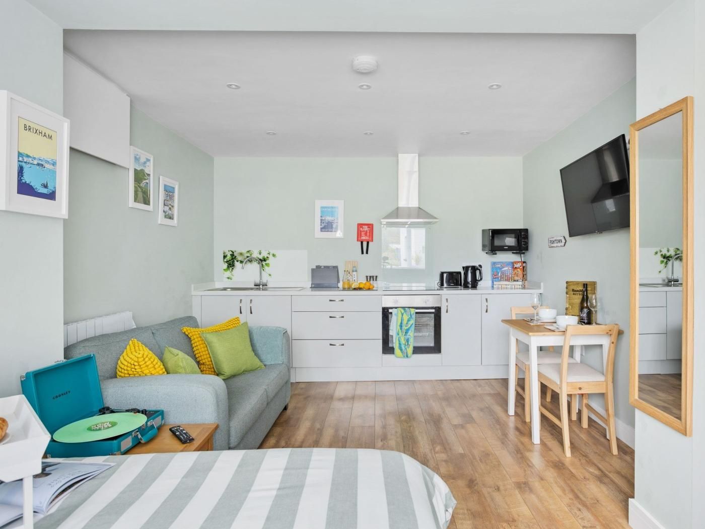 Ein gemütlicher Wohnraum mit einer Küchenzeile, einem Sofa mit bunten Kissen, einem Essbereich mit Tisch und Stühlen sowie einem an der Wand befestigten Fernseher.