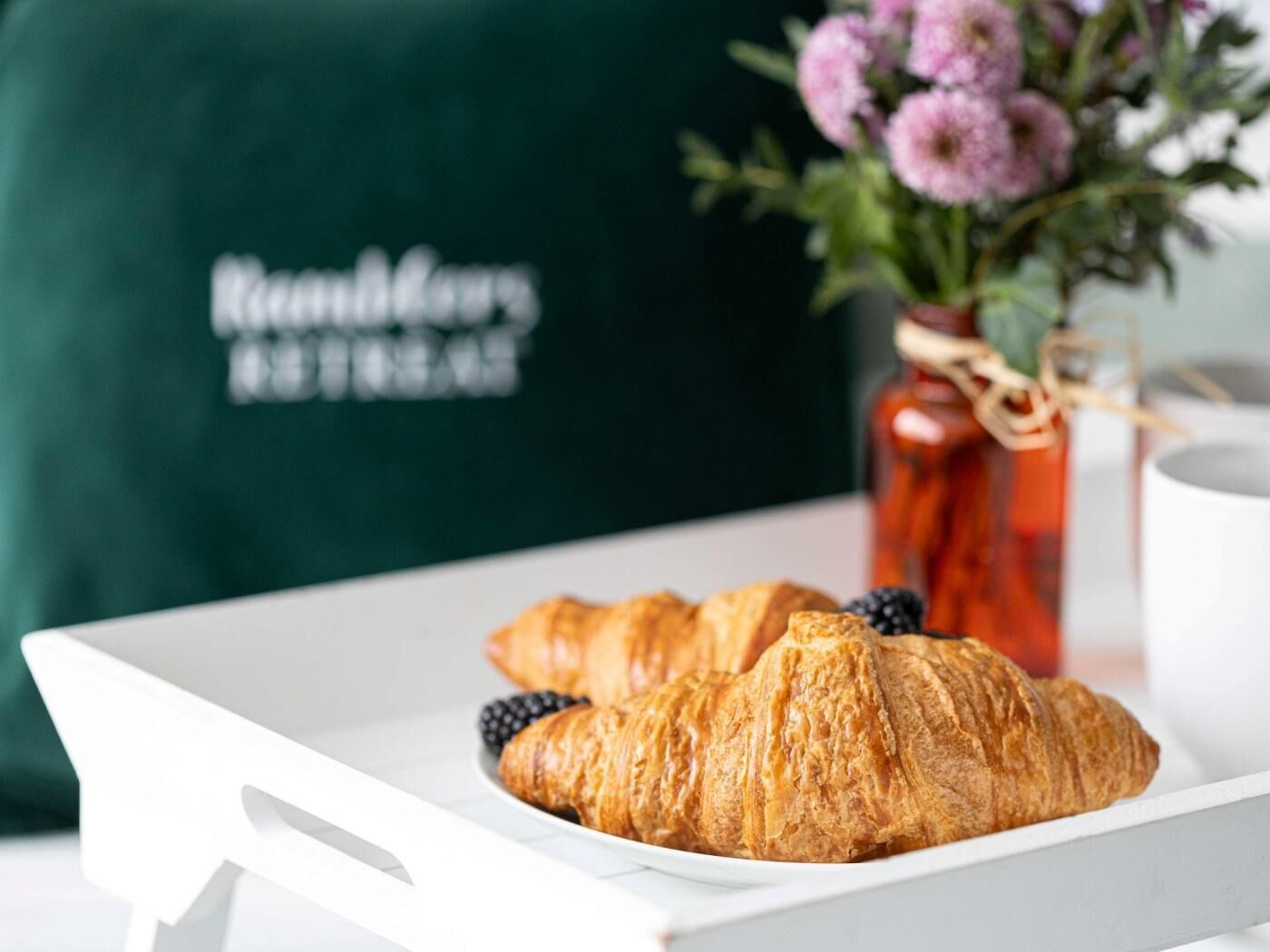 A tray with croissants and blackberries on a white tray, a green apron with text, and a vase with flowers in the background.