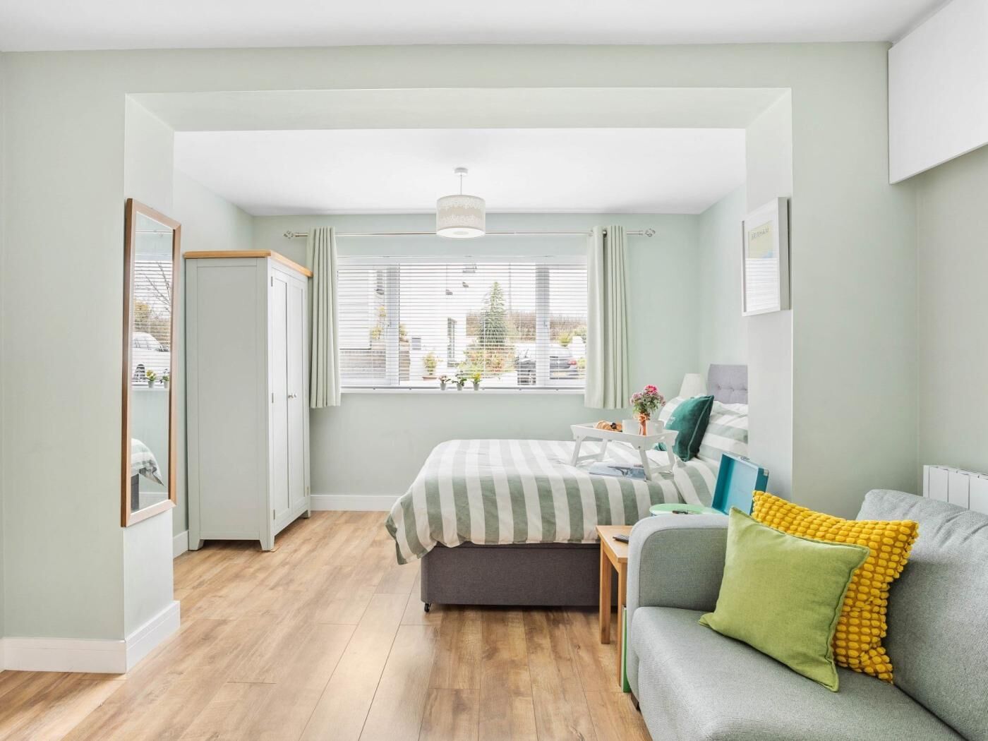 A bedroom with a bed, a sofa, a window with blinds, and a door.