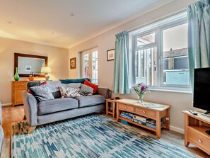 Ein Wohnzimmer mit grauem Sofa, einem Holztisch, einem Fernsehschrank, einem Fenster mit Vorhängen und einem gemusterten Teppich.