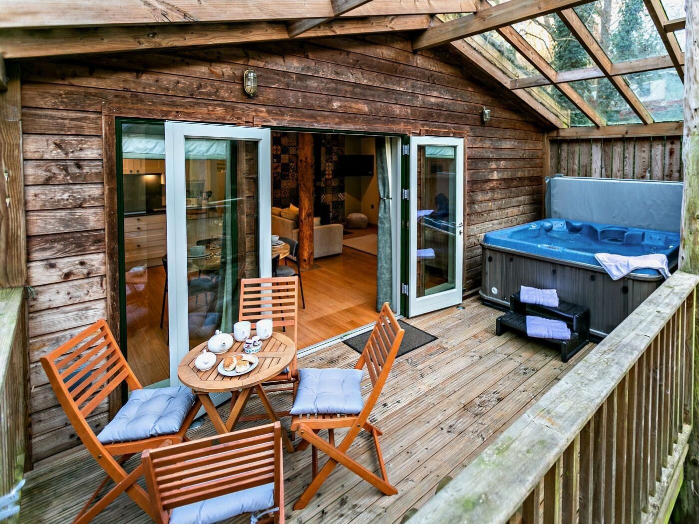 A wooden deck with a hot tub, a dining area with a table and chairs, and a sliding glass door leading to an interior space.