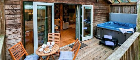 A wooden deck with a hot tub, a dining area with a table and chairs, and a sliding glass door leading to an interior space.