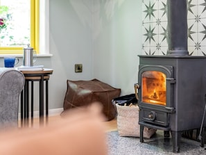 A cozy room with a wood-burning stove, a window with a view, a small table with a kettle, and a chair. - 3 Bed in Truro (oc-93308) (Porthtowan)