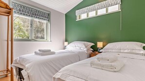 A hotel room with two beds, a green accent wall, a window with patterned curtains, and a wooden floor. - 2 Bed in Truro (oc-93310) (Porthtowan)