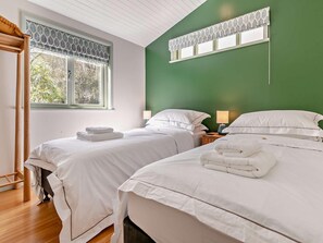 A hotel room with two beds, a green accent wall, a window with patterned curtains, and a wooden floor. - 2 Bed in Truro (oc-93310) (Porthtowan)