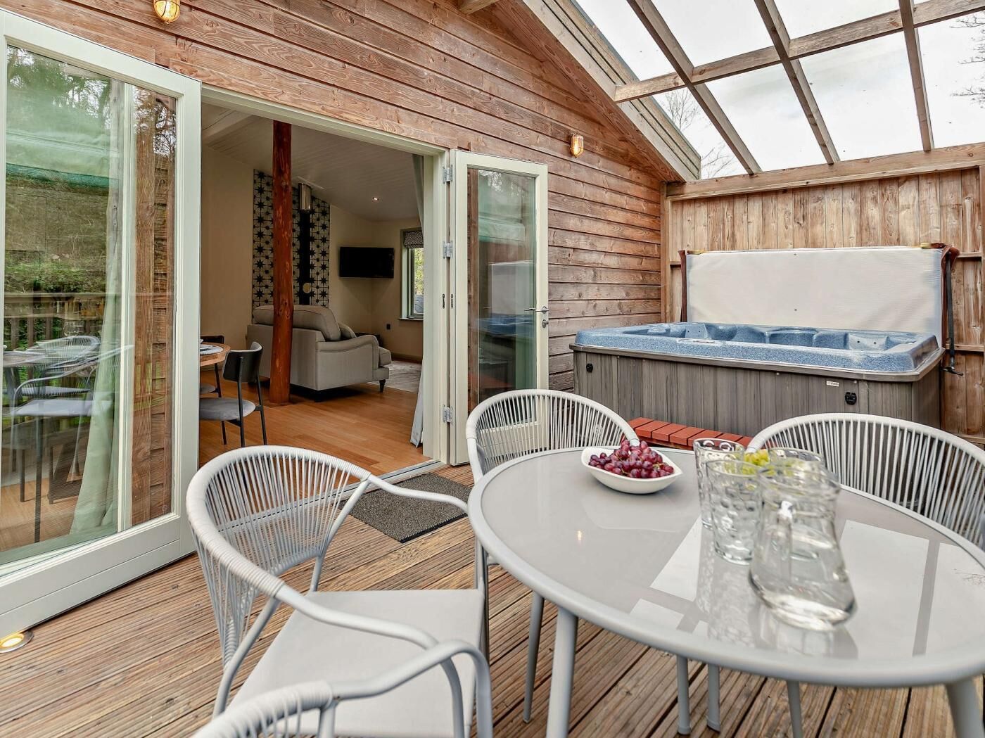 En træterrasse med en glasdør, der fører til et opholdsområde indendørs, et spabad og et bord med en skål frugt.