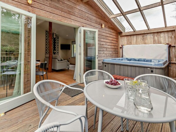 A wooden deck with a glass door leading to an interior living area, a hot tub, and a table with a bowl of fruit. - 2 Bed in Truro (oc-93310) (Porthtowan)