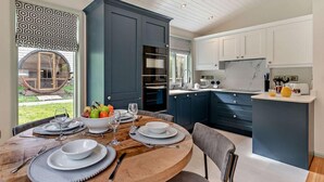 A modern kitchen with dark blue cabinets, a wooden dining table set with plates and glasses, and a view of the outdoors through a large window. - 2 Bed in Truro (oc-93310) (Porthtowan)