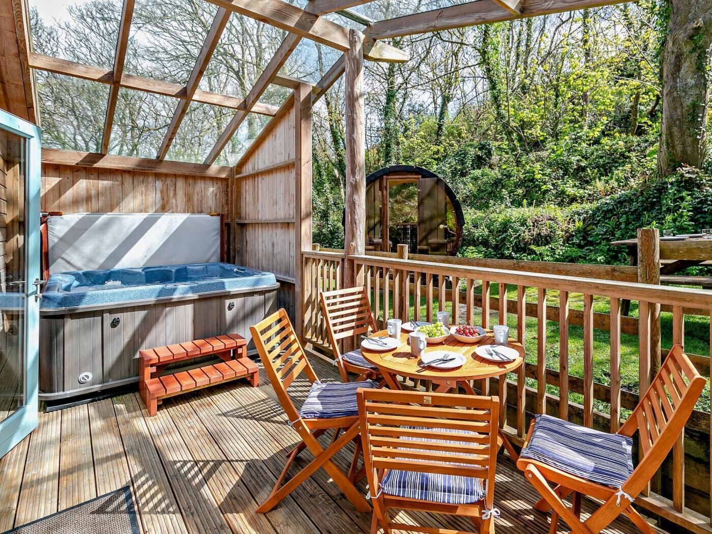 Eine Holzterrasse mit Whirlpool, Sitzgelegenheiten im Freien und Blick auf die umliegende Grünfläche.